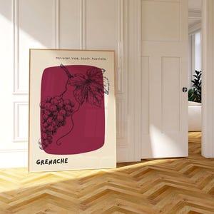 May include: A framed poster featuring a wine-themed illustration. The artwork depicts a bunch of grapes and a leaf in black ink against a maroon background. The text "Grenache" is at the bottom, with "McLaren Vale, South Australia" at the top.