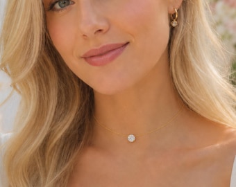 May include: A delicate gold chain necklace featuring a single round clear stone pendant, worn with small gold hoop earrings. The jewelry is paired with a white short-sleeved top with puffed sleeves and a square neckline, creating a clean look.