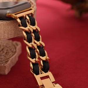 May include: Gold-toned watch band with a black woven chain design. The band features a gold clasp and connectors, creating a luxurious look. The watch band is shown against a red background.