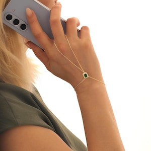 May include: A delicate gold hand chain bracelet. The bracelet features a small, square, emerald-colored gemstone with a halo of small, clear stones. The chain extends from the bracelet to a ring that is worn on the middle finger.