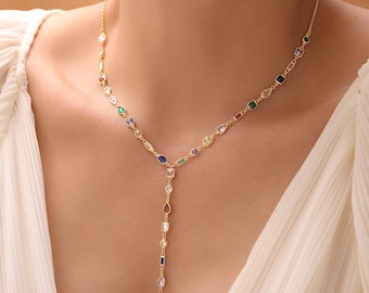 May include: A gold-toned necklace with a Y-shaped pendant. The necklace features a delicate chain adorned with a variety of colorful, geometric gemstones, including emerald green, sapphire blue, and ruby red. The pendant is a vertical line of gemstones.