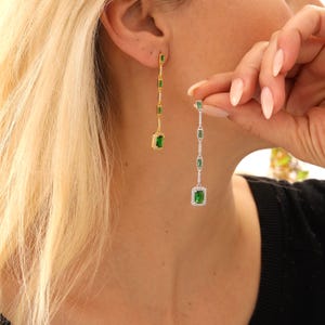 May include: A pair of dangling earrings with emerald-cut green gemstones. One earring is gold-toned, the other is silver-toned. The earrings are rectangular with smaller gemstones and a larger square gemstone at the bottom. The earrings are being held up to the ear.