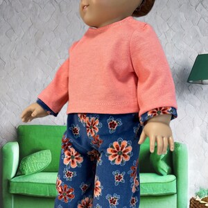 May include: A pink long-sleeved shirt and blue pants with a floral print. The pants have a red, orange, and white floral pattern. The shirt has a crew neck.