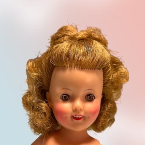 May include: A vintage doll with blonde, curly hair styled in a bouffant. The doll has a fair complexion, rosy cheeks, and red lipstick. The doll's eyes are brown and wide, and it has a slight smile. The doll is a collectible item.