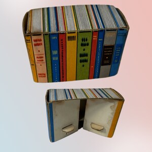K&ouml;nnte beinhalten: Zwei Pappschachteln, eine &uuml;ber der anderen, die wie ein Stapel B&uuml;cher aussehen sollen. Die Schachteln sind rechteckig und haben farbenfrohe Buchr&uuml;cken auf den Seiten. Die obere Schachtel ist geschlossen, die untere Schachtel ist offen.