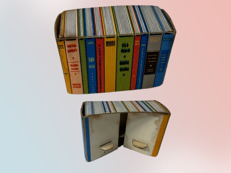 K&ouml;nnte beinhalten: Zwei Pappschachteln, eine &uuml;ber der anderen, die wie ein Stapel B&uuml;cher aussehen sollen. Die Schachteln sind rechteckig und haben farbenfrohe Buchr&uuml;cken auf den Seiten. Die obere Schachtel ist geschlossen, die untere Schachtel ist offen.