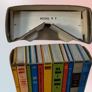 K&ouml;nnte beinhalten: Eine Vintage-Kartonbox, die wie ein Stapel B&uuml;cher gestaltet ist. Die Box ist mit farbenfrohen Buchr&uuml;cken in verschiedenen Farben verziert. Die Oberseite der Box ist offen und zeigt den Text "BOOKS B 2" auf einer wei&szlig;en Karte.