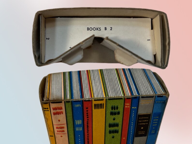 K&ouml;nnte beinhalten: Eine Vintage-Kartonbox, die wie ein Stapel B&uuml;cher gestaltet ist. Die Box ist mit farbenfrohen Buchr&uuml;cken in verschiedenen Farben verziert. Die Oberseite der Box ist offen und zeigt den Text "BOOKS B 2" auf einer wei&szlig;en Karte.