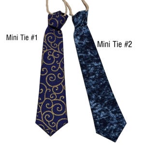May include: Two mini ties are displayed against a white background. One is navy blue with gold swirls, labeled "Mini Tie #1." The other is a dark blue, textured pattern, labeled "Mini Tie #2." Both ties have a beige string attached.