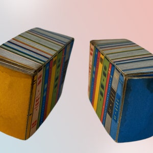 K&ouml;nnte beinhalten: Zwei dekorative, buchf&ouml;rmige Schachteln. Jede Schachtel ist so gestaltet, dass sie einem Stapel B&uuml;cher &auml;hnelt, mit farbenfrohen Buchr&uuml;cken und Titeln. Eine Schachtel hat eine gelbe Seite, die andere ist blau. Die Oberseiten der Schachteln sind silberfarben.