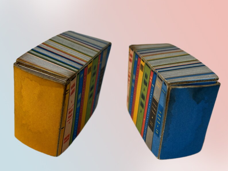 K&ouml;nnte beinhalten: Zwei dekorative, buchf&ouml;rmige Schachteln. Jede Schachtel ist so gestaltet, dass sie einem Stapel B&uuml;cher &auml;hnelt, mit farbenfrohen Buchr&uuml;cken und Titeln. Eine Schachtel hat eine gelbe Seite, die andere ist blau. Die Oberseiten der Schachteln sind silberfarben.