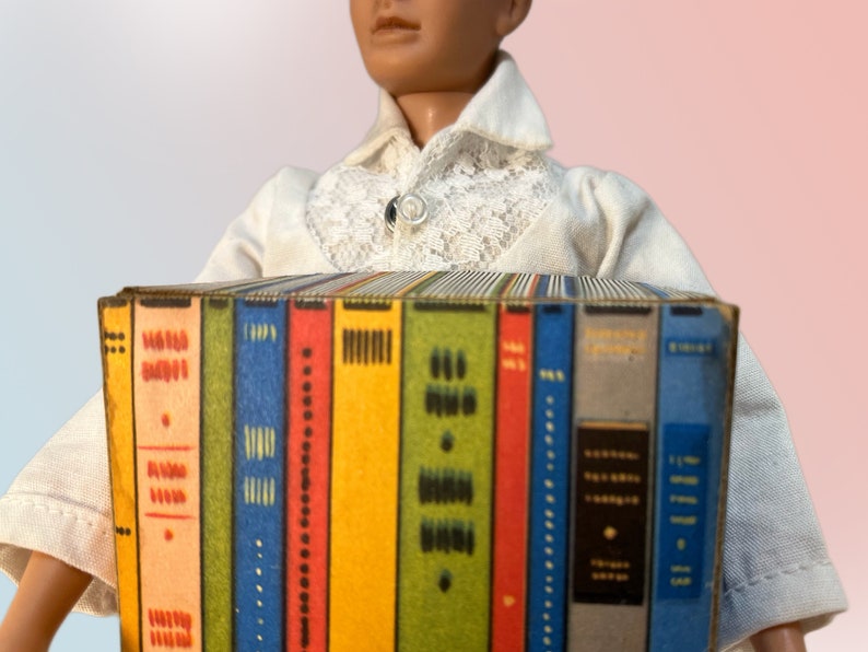K&ouml;nnte beinhalten: Eine Sammlung von Miniaturb&uuml;chern mit farbenfrohen Buchr&uuml;cken. Die B&uuml;cher werden von einer Puppe gehalten, die ein wei&szlig;es Hemd mit Spitzenkragen tr&auml;gt. Die Buchr&uuml;cken sind in verschiedenen Farben gehalten, darunter Rot, Blau, Gr&uuml;n und Gelb.