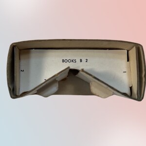 K&ouml;nnte beinhalten: Ge&ouml;ffnete, alte Pappschachtel mit dem Text "BOOKS B 2" auf einer wei&szlig;en Innenfl&auml;che. Die Schachtel weist Gebrauchsspuren auf, mit Knicken und Rissen, was auf ein antikes Objekt hindeutet.