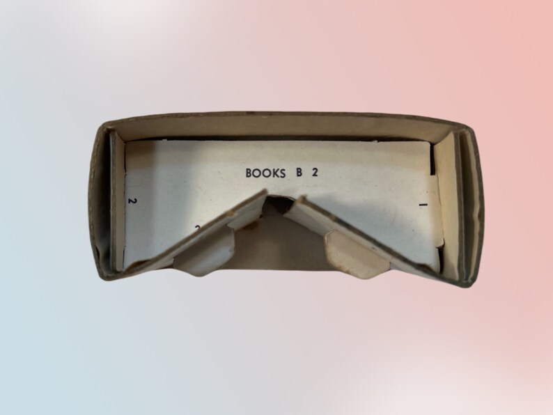 K&ouml;nnte beinhalten: Ge&ouml;ffnete, alte Pappschachtel mit dem Text "BOOKS B 2" auf einer wei&szlig;en Innenfl&auml;che. Die Schachtel weist Gebrauchsspuren auf, mit Knicken und Rissen, was auf ein antikes Objekt hindeutet.