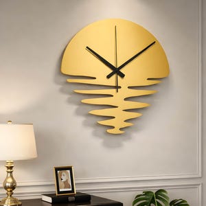 May include: A gold-colored wall clock with a unique design resembling a sunset. The clock features black hands and a stylized sun shape with wavy lines below, creating a modern and artistic aesthetic. The clock is mounted on a light gray wall.