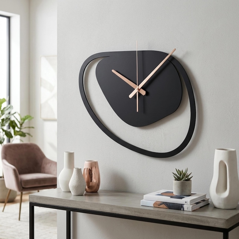 Pu&ograve; includere: Un moderno orologio da parete nero con lancette color oro rosa. L'orologio ha un design astratto con una forma ovale nera e un anello nero circostante. &Egrave; montato su una parete bianca, con vasi decorativi e libri su un tavolo sottostante.