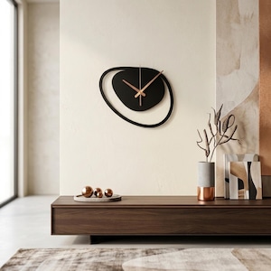 Pu&ograve; includere: Un moderno orologio da parete nero con un design astratto unico. L'orologio presenta una forma ovale nera con un cerchio interno nero pi&ugrave; piccolo e lancette color rame. Montato su una parete color crema, sopra un mobile in legno scuro. Misura circa 40 cm di diametro.