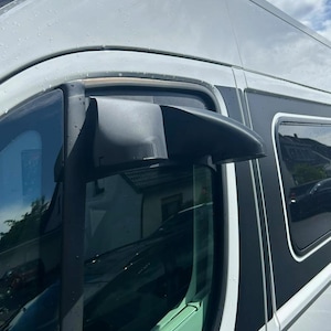 Könnte beinhalten: Ein schwarzer Windabweiser oder Regenschutz aus Kunststoff, der am oberen Rahmen einer weißen Transportertür montiert ist. Das Zubehörteil hat eine geschwungene, aerodynamische Form, die über dem Seitenfenster angebracht ist.