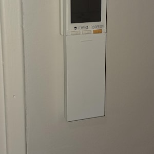 Może przedstawiać: Biały pilot do klimatyzacji Mitsubishi Electric zamontowany na białej ścianie. Pilot posiada czarny ekran oraz przyciski z napisami "TEMP" i "OFF/ON". Logo Mitsubishi jest widoczne u góry.