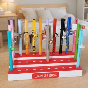 Può includere: Un portapenne rosso e bianco con una varietà di penne colorate. Le penne sono decorate con ciondoli a tema animale. Il portapenne ha il testo "LEGAMI DE PRENOM" sulla parte anteriore.