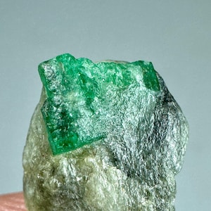 May include: A raw emerald crystal with a vibrant green hue, set against a matrix of gray and tan rock. The emerald's crystalline structure is visible, showcasing its natural, unpolished form. The stone is a focal point, with a rough, textured appearance.