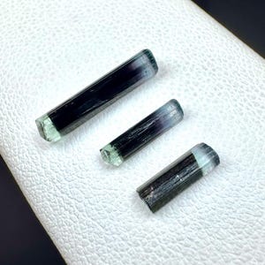 May include: Three tourmaline crystals with a dark green and black color gradient. The crystals are cylindrical with a faceted, light green end. The crystals are arranged diagonally on a white, textured surface.