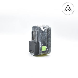 May include: A used, weathered black and gray power tool battery with a black base and a small green sticker. The battery is rectangular with a ridged design on the side. The background is white.
