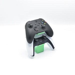 Pode incluir: Um comando de jogo sem fios preto repousa sobre um comando de jogo sem fios branco e preto, ambos apoiados por um suporte de plástico verde claro. Os comandos apresentam vários botões, joysticks e painéis direcionais.