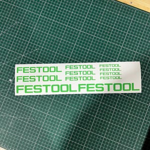 Op de afbeelding: Een witte sticker met het woord "FESTOOL" in het groen meerdere keren afgedrukt. De sticker ligt op een groene snijmat met een rasterpatroon.