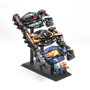 Op de afbeelding: Een display van drie Formule 1-raceauto's gemaakt van zwarte, oranje, blauwe en witte bouwstenen. De auto's zijn gemonteerd op een zwarte standaard, met drie miniatuurfiguren aan de basis.