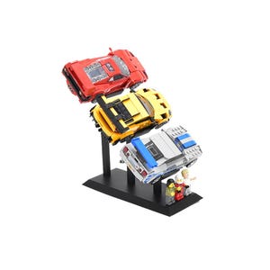 Op de afbeelding: Een zwarte displaystandaard met drie speelgoedauto's in rood, geel en wit met blauwe strepen. De auto's zijn gemonteerd op zwarte steunen, met kleine Lego-figuren aan de basis. De auto's staan schuin om te lijken alsof ze in beweging zijn.