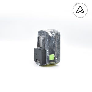 May include: A used, weathered black and gray power tool battery with a black base and a small green sticker. The battery has a rectangular shape with a ridged top and is set against a white background.