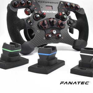 Op de afbeelding: Een zwart en grijs Fanatec racestuur met meerdere knoppen en draaiknoppen. Het stuur heeft een koolstofvezel ontwerp en een suède-achtige grip. Drie zwarte stands met gekleurde accenten staan op de voorgrond.