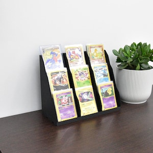 May include: A black tiered display stand holding eight trading cards in clear protective sleeves. The stand features three rows arranged in a stepped design, showcasing various colorful character illustrations and text on each individual card.