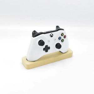 Puede incluir: Un mando Xbox blanco con detalles negros, apoyado en un soporte marrón claro. El mando tiene botones de colores y una cruceta. El soporte es rectangular con bordes redondeados. El fondo es blanco.