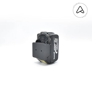 May include: A black Makita 18V 3.0Ah Lithium-Ion battery pack with a black mounting bracket. The battery pack has white text that reads "18V" and "3.0Ah Lithium Ion". The battery is shown against a white background.