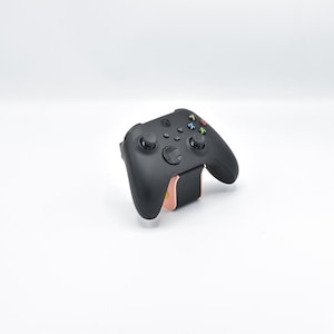 Pode incluir: Um comando de videojogos sem fios preto com dois joysticks analógicos, um pad direcional e quatro botões de ação coloridos com as etiquetas X, Y, A e B. O comando está montado num pequeno suporte cor de pêssego na posição vertical.