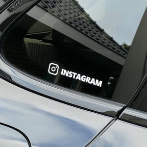 May include: White Instagram logo and text sticker on a car window. The Instagram logo is a white square with a white circle and a white dot inside. The word "INSTAGRAM" is written in white, sans-serif font.