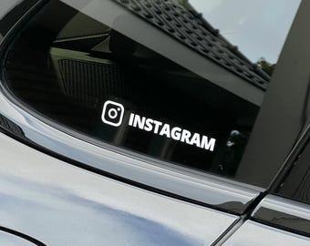 May include: White Instagram logo and text decal on a car window. The Instagram logo is a white square with a white circle and a white dot inside. The word "INSTAGRAM" is written in white, sans-serif font.