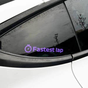 May include: A purple sticker on a car window displaying the text "Fastest lap" and a stopwatch symbol. The sticker is affixed to a white car with black detailing.