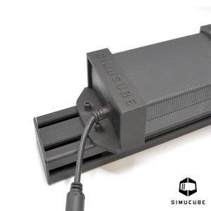 Könnte beinhalten: Eine schwarze Simucube-Elektronikkomponente, die auf einer schwarzen Aluminium-Extrusionsschiene montiert ist. Das Gerät verfügt über ein strukturiertes rechteckiges Gehäuse mit dem geprägten Text SIMUCUBE und einem schwarzen Stromkabel.