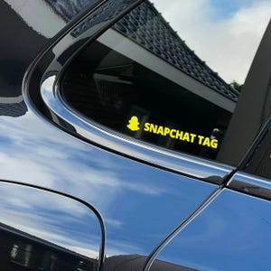 May include: A yellow Snapchat tag sticker is affixed to the window of a black car. The sticker features the Snapchat ghost logo and the words "SNAPCHAT TAG" in a bold, sans-serif font. The car's glossy surface reflects the sky.