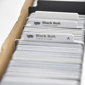 Pode incluir: Uma caixa de cartas colecionáveis com divisórias brancas com a etiqueta "Black Bolt Scarlet & Violet". As cartas são maioritariamente cinzentas com bordas brancas. A caixa é de cartão.