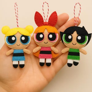 Puede incluir: Tres adornos de fieltro que representan a los personajes de Las Supernenas: Burbuja, Pétalo y Cactus. Cada uno tiene ojos grandes, una cara sonriente y un peinado único. Visten sus trajes característicos y tienen una cuerda roja y blanca para colgar.