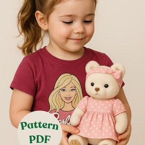 Peut inclure: Un ours en peluche vêtu d'une robe rose à pois et d'un bandeau assorti. L'ours a un nez et des pattes bruns. L'enfant porte un t-shirt bordeaux avec un motif Barbie et le texte "Pattern PDF".