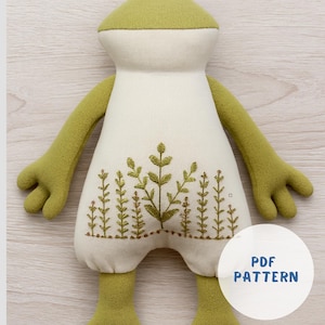 Può includere: Un giocattolo a forma di rana di peluche fatto a mano con testa, braccia e gambe verde chiaro e un corpo color crema. Il corpo presenta un design botanico ricamato. L'immagine include un cerchio bianco con il testo "PDF PATTERN".