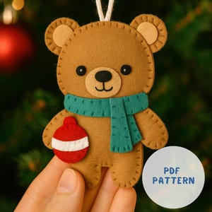 May include: A brown felt teddy bear Christmas ornament with a green scarf and a red and white ornament. The bear has black eyes and a black nose. The ornament is held by a white string. The text "PDF PATTERN" is on a white circle.