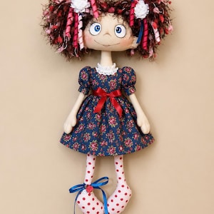 Pode incluir: Uma boneca de pano com cabelo castanho escuro texturizado, detalhes florais brancos e olhos azuis. A boneca veste um vestido azul-marinho com estampa floral vermelha, gola de renda branca e laço vermelho na cintura. As pernas têm bolinhas vermelhas.