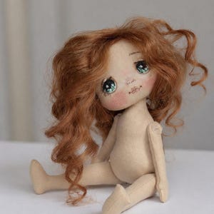 May include: A handcrafted fabric doll with a beige body, featuring long, wavy, copper-colored hair. The doll has large, detailed green eyes with visible eyelashes, rosy pink cheeks, and a small stitched mouth, sitting in a seated position.