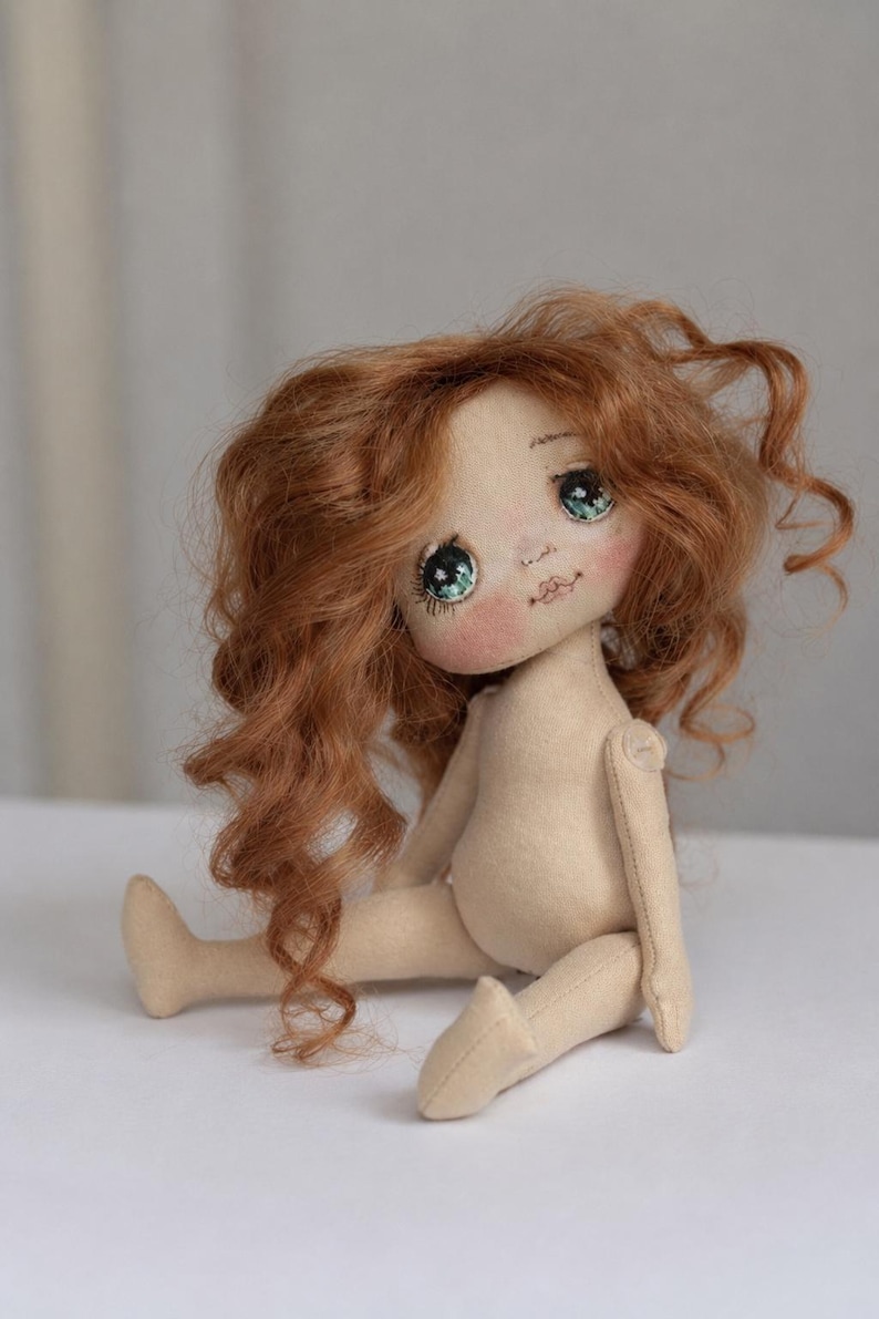 May include: A handcrafted fabric doll with a beige body, featuring long, wavy, copper-colored hair. The doll has large, detailed green eyes with visible eyelashes, rosy pink cheeks, and a small stitched mouth, sitting in a seated position.